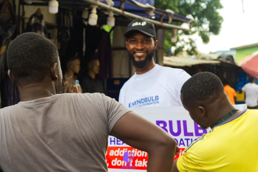 A smiling volunteer holding the Mindbulb banner while speaking with two young men at a market